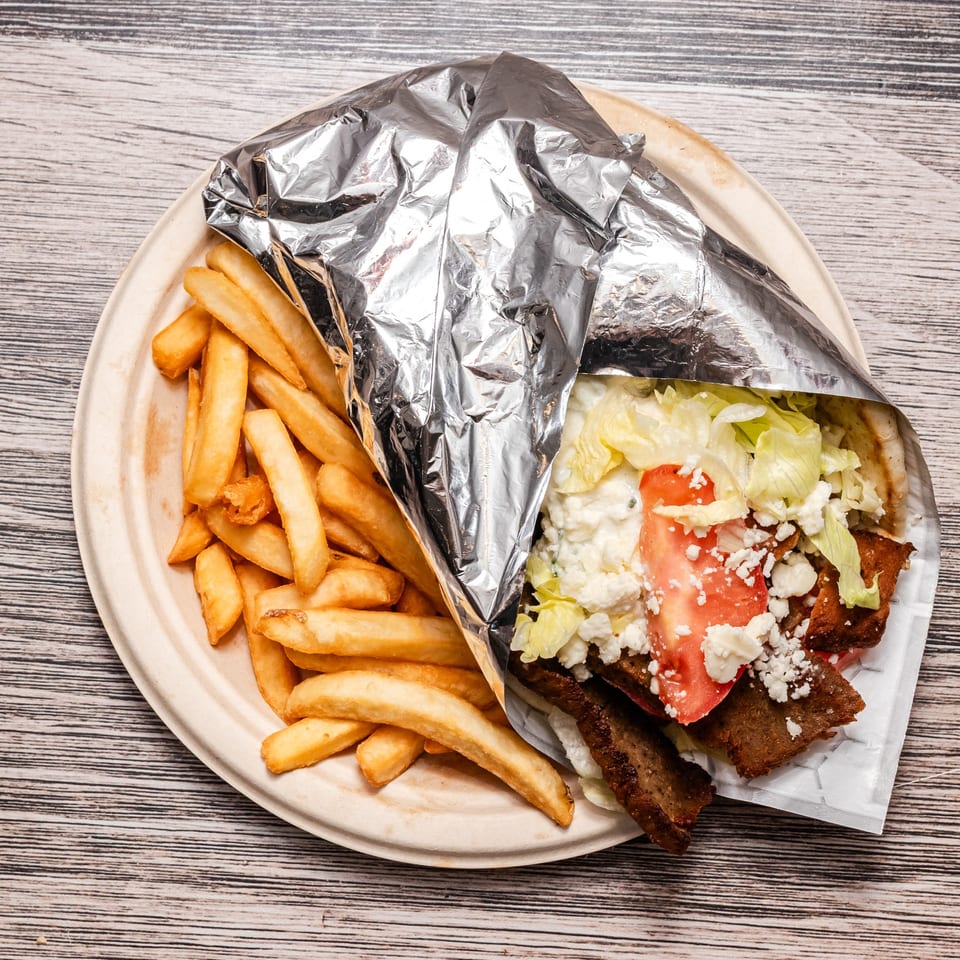 Gyro pita wrap with lettuce, tomato, feta, and fries on a paper plate, partially wrapped in foil.