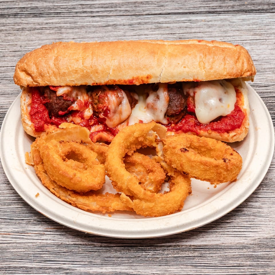 Meatball sub with marinara and melted cheese served alongside crispy onion rings on a plate.