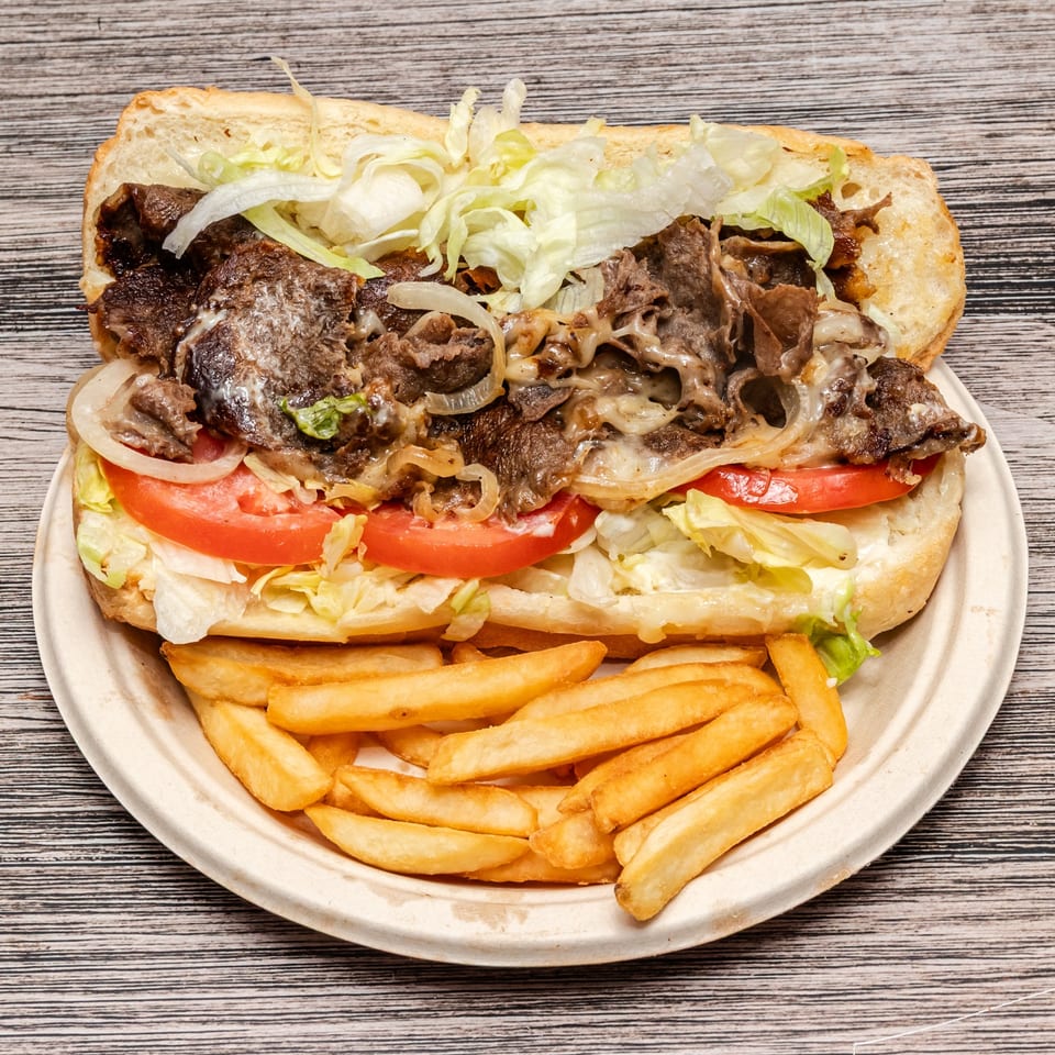 Loaded steak sandwich with melted cheese, onions, lettuce, and tomatoes served alongside golden French fries on a plate.