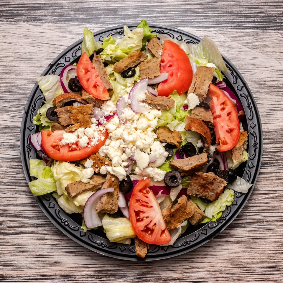 Greek salad with lettuce, tomatoes, red onions, black olives, feta cheese, and sliced seasoned meat on a black platter.